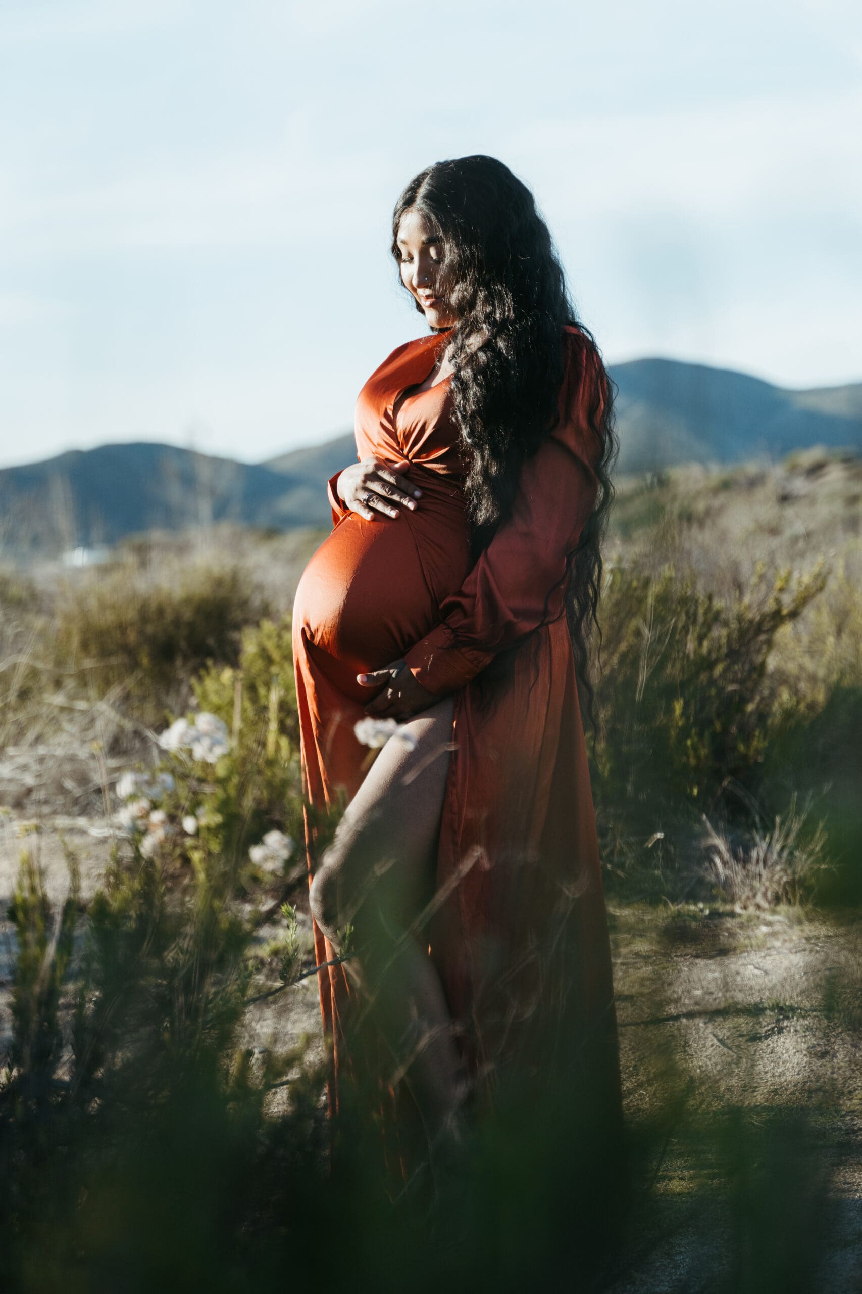 Image of a glowing pregnant woman in a orange gown, standing in a golden field at sunset, cradling her bump and smiling peacefully.
