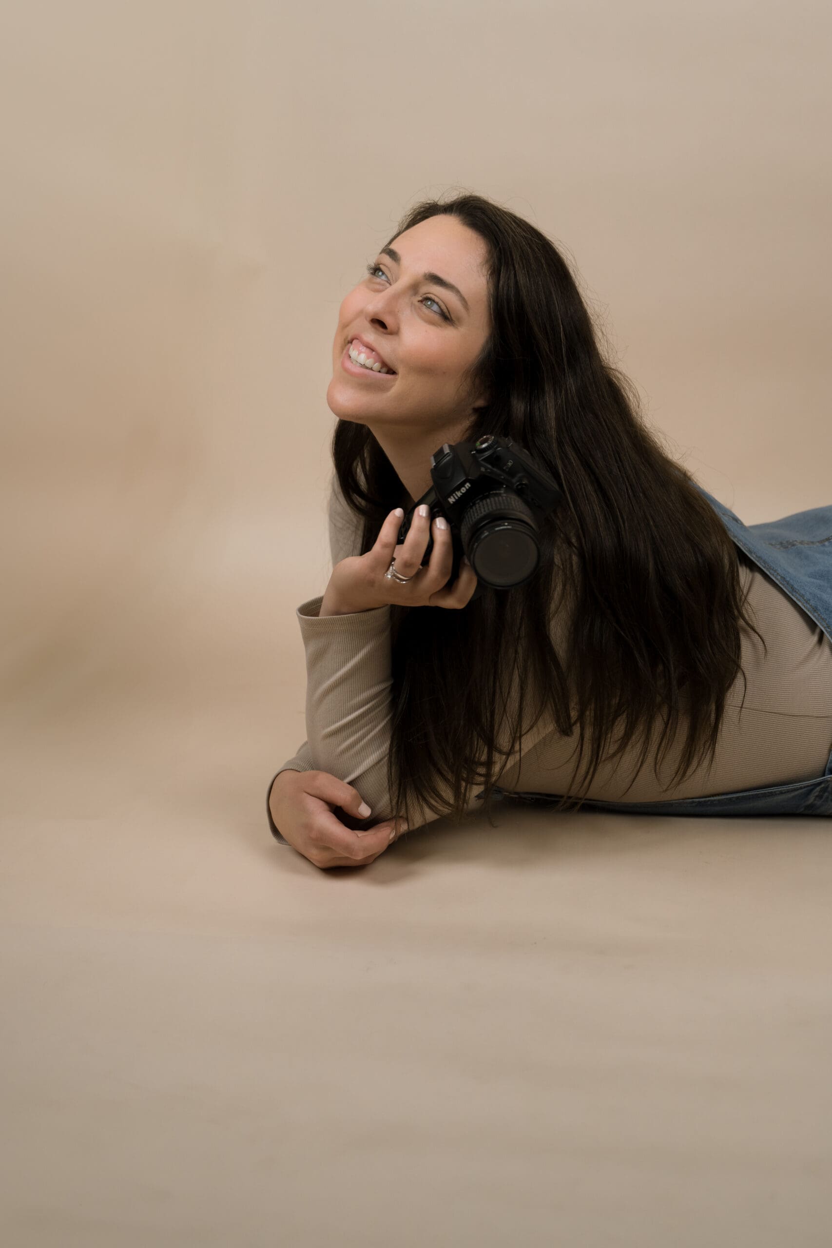 Image of a brunette laying down in overalls holding her Nikon while looking off in the distance.